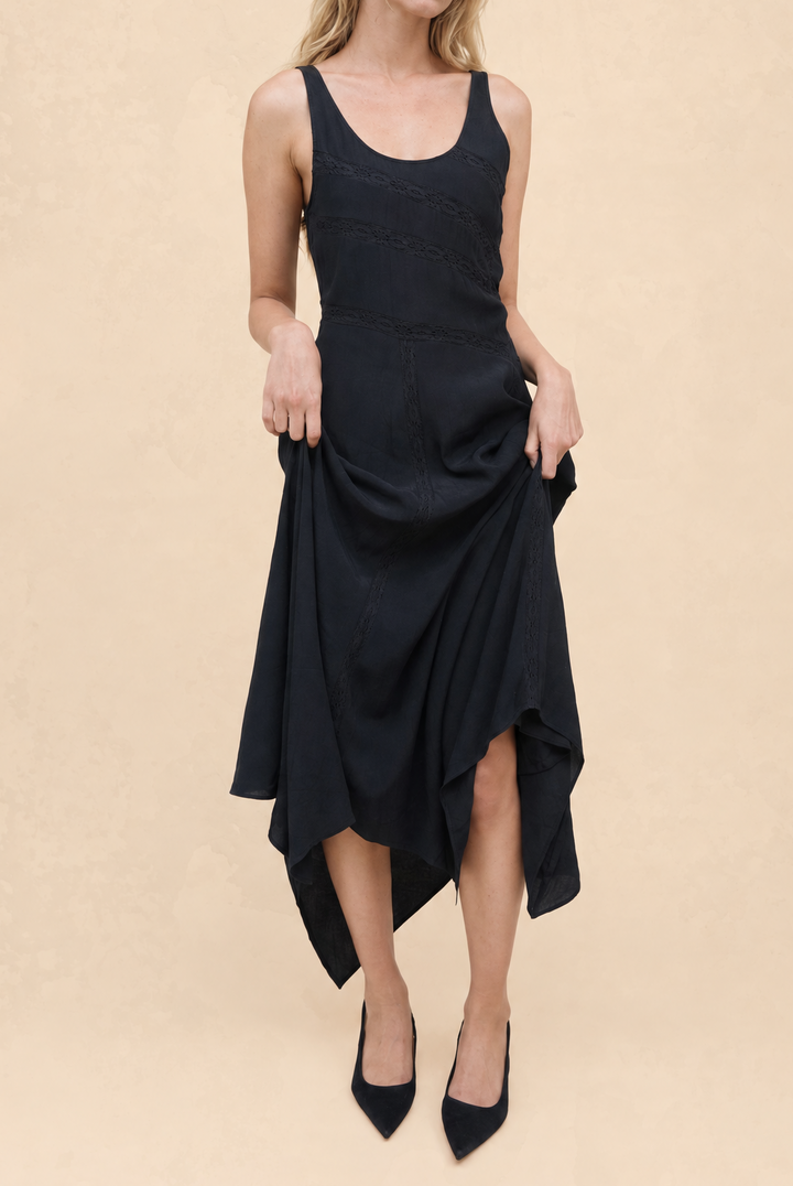 A woman modeling a sleeveless black midi dress with a scoop neckline, textured bodice, and draped asymmetrical skirt, paired with black pointed-toe heels against a warm beige studio background.
