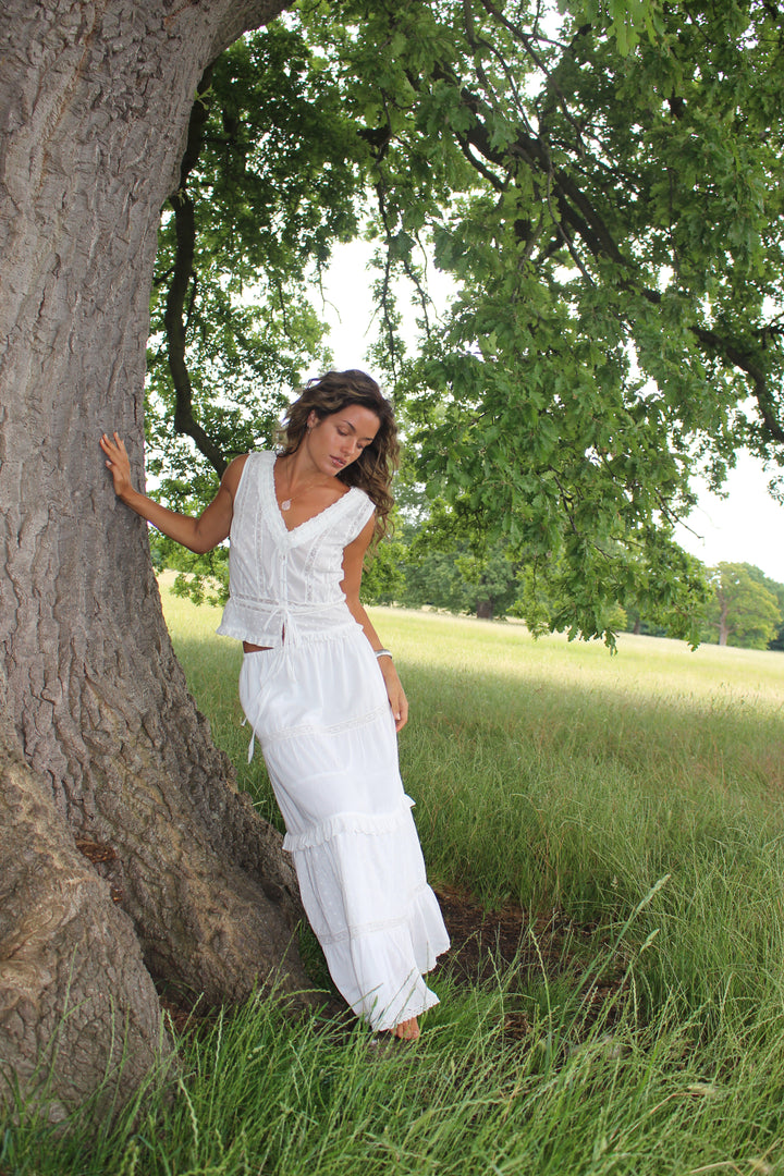 The Violetta Top and Skirt in white cotton lace-trim top with deep V neckline and tie-front detail, shot in greenery. 