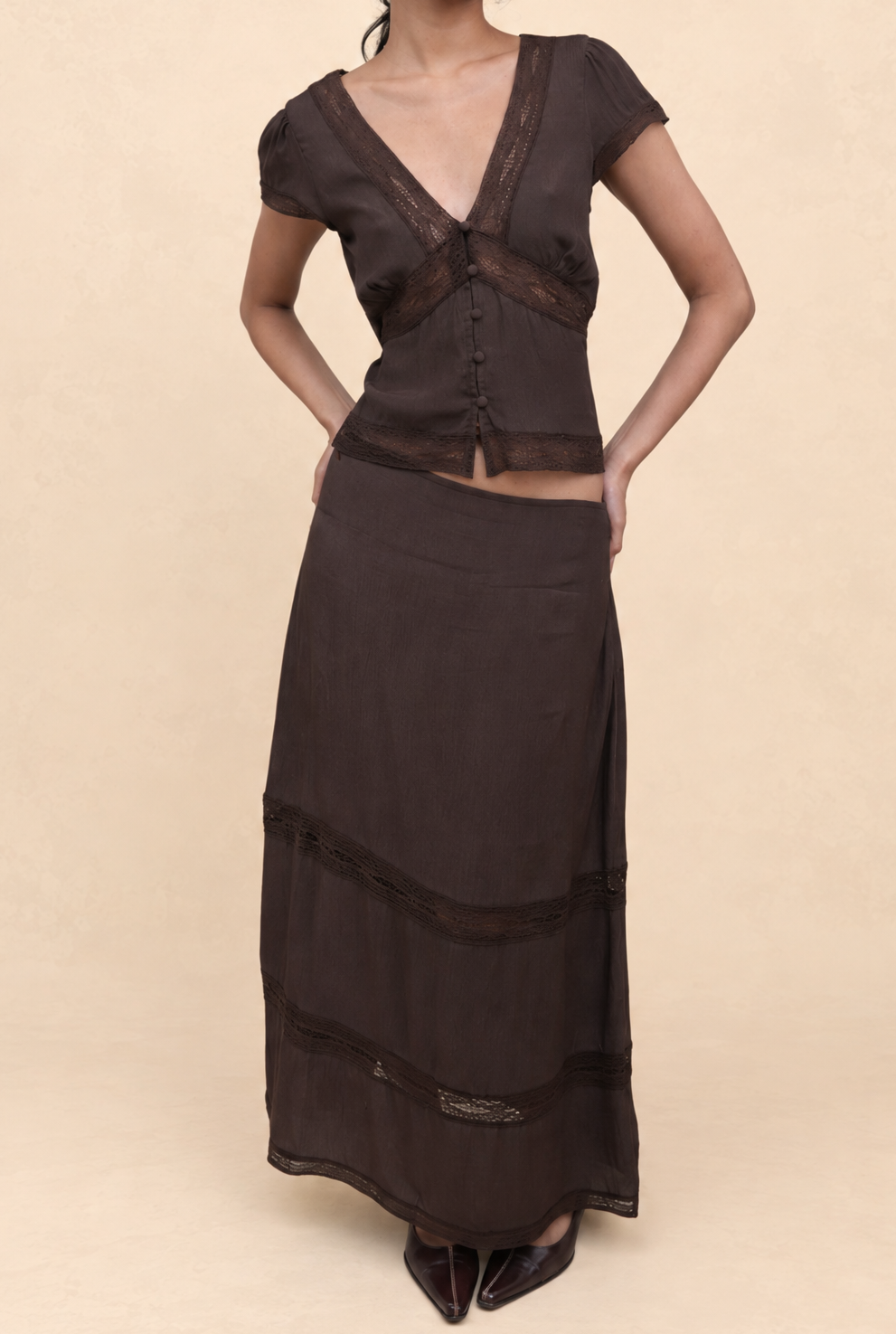 A woman modeling a dark brown two-piece outfit consisting of a short-sleeve V-neck top with sheer mesh panels and a matching long skirt, set against a solid beige studio background.