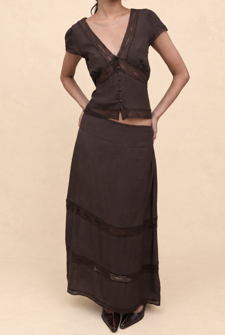 A woman modeling a dark brown two-piece outfit consisting of a short-sleeve V-neck top with sheer mesh panels and a matching long skirt, set against a solid beige studio background.