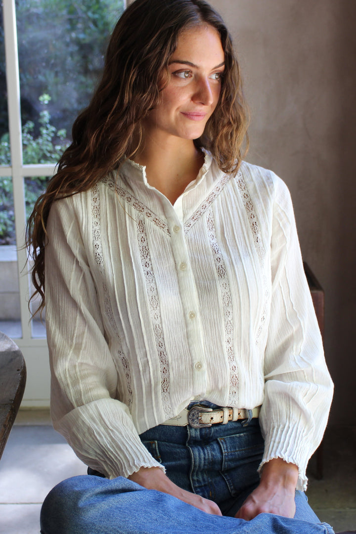 A woman wearing a cream-colored, long-sleeved button-up blouse with vertical lace or embroidered stripes, paired with blue jeans, sitting in a sunlit room.