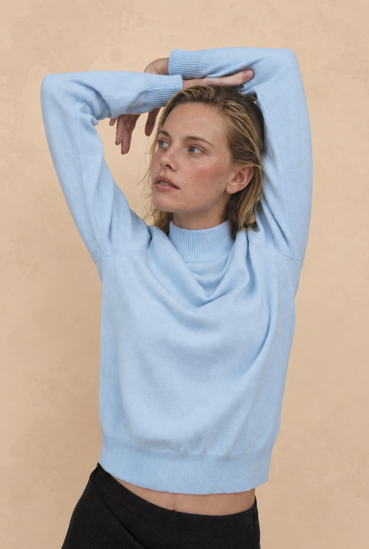 A woman wearing a light blue mock-neck knit sweater and a black skirt, posing with her arms raised against a textured beige background.