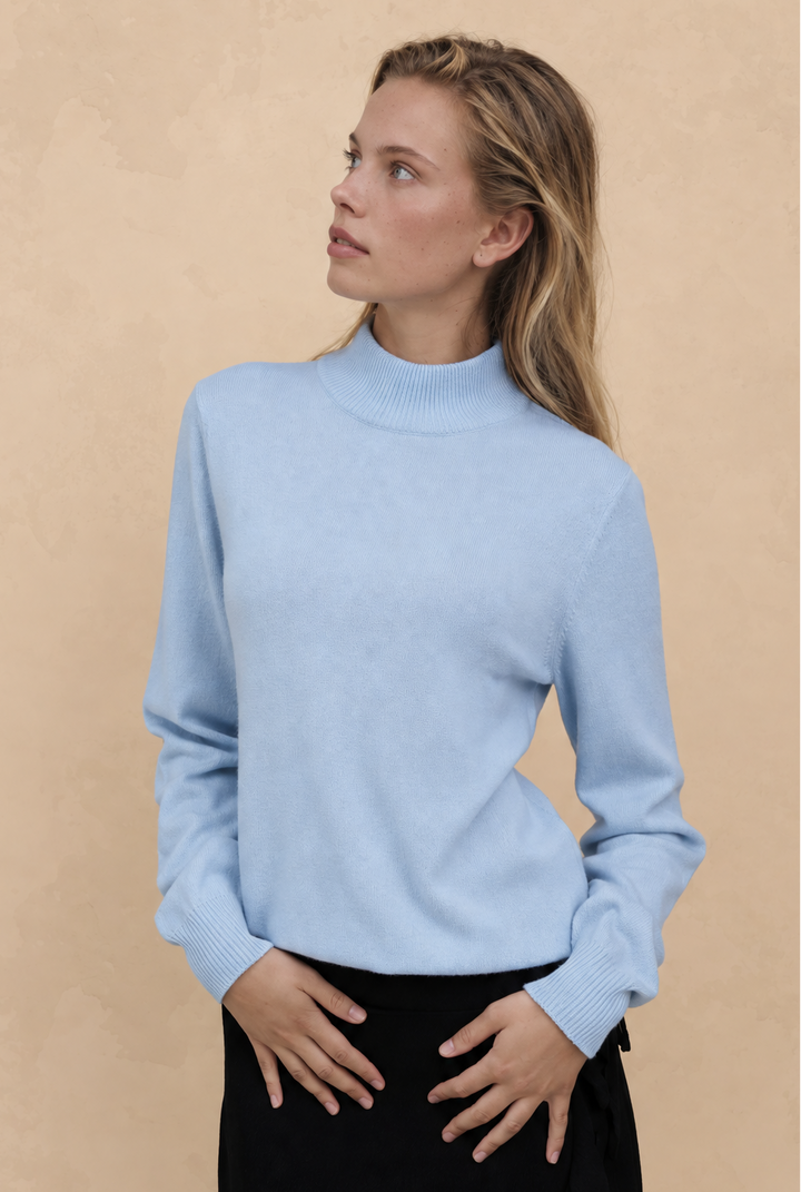 A woman wearing a light blue mock neck knit sweater and a black skirt, standing against a textured beige background.