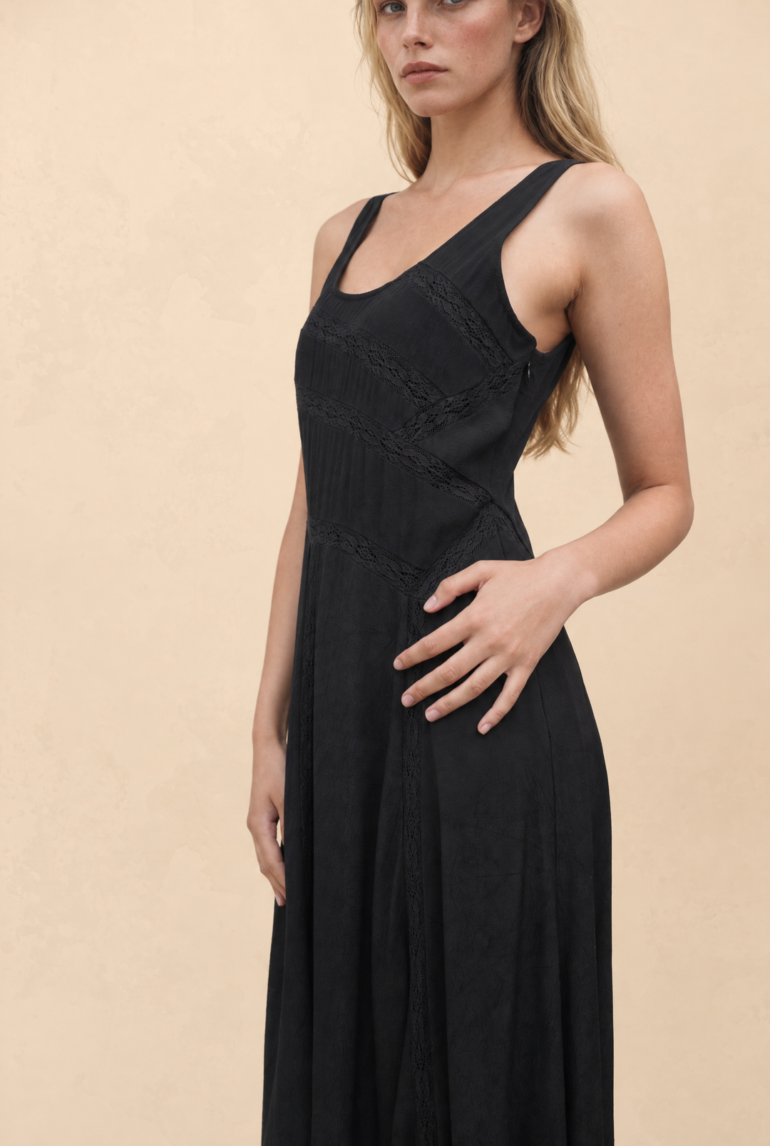 A woman wearing a sleeveless black maxi dress with a scoop neckline, lace-up front, and gathered waist details, posed against a warm beige textured background.