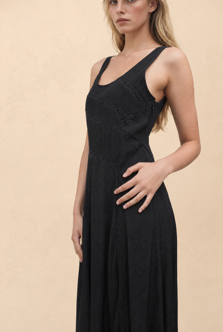 A woman wearing a sleeveless black maxi dress with a scoop neckline, lace-up front, and gathered waist details, posed against a warm beige textured background.